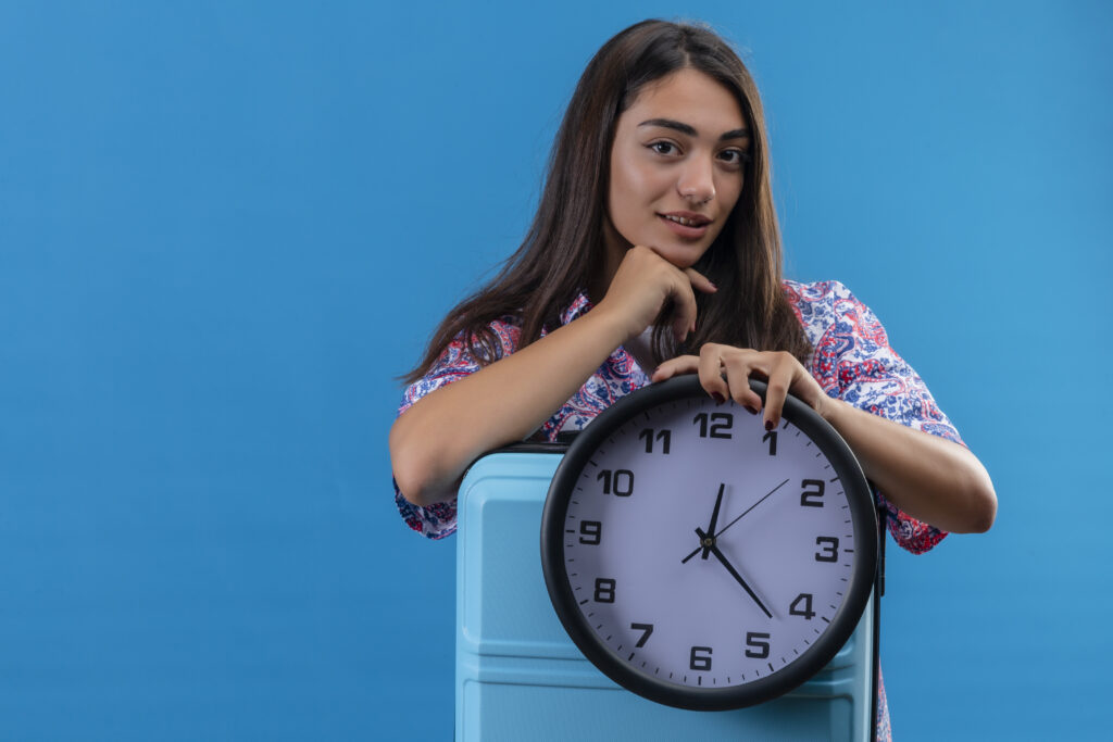 young beautiful traveler woman holding blue suitcase round clock with hand chin thinking waiting blue wall