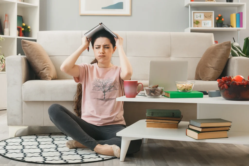 unpleased with closed eyes young girl covered head with notebook sitting floor coffee table living room
