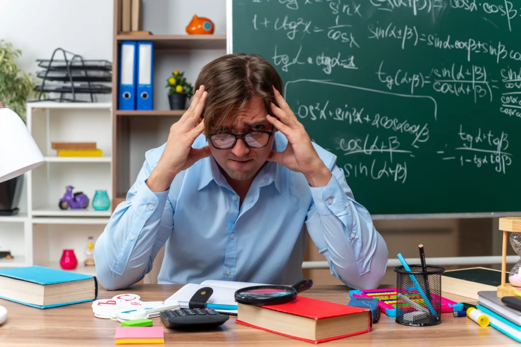 young male teacher wearing glasses stressed nervous sitting school desk with books notes front blackboard classroom